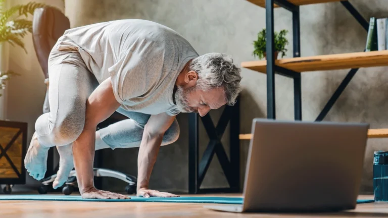 Older Male Patient Performing Yoga To Stretch Back In Front Of A Laptop